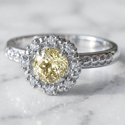 A light yellow round Chatham sapphire engagement ring with a halo of round gold beads and lab grown diamonds set in a white gold band.