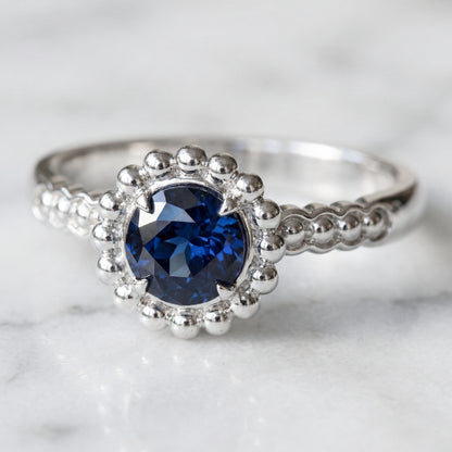 A round medium blue sapphire engagement ring with a halo of gold spheres set in a white gold band.