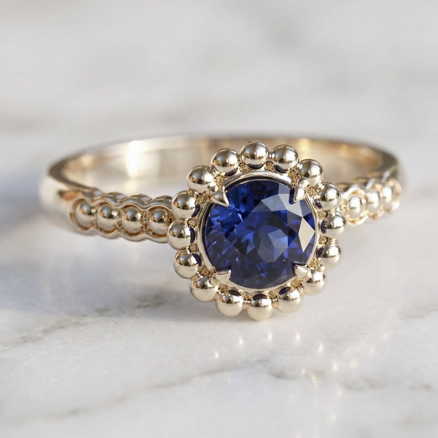 A round medium blue sapphire engagement ring with a halo of gold spheres set in a gold band.