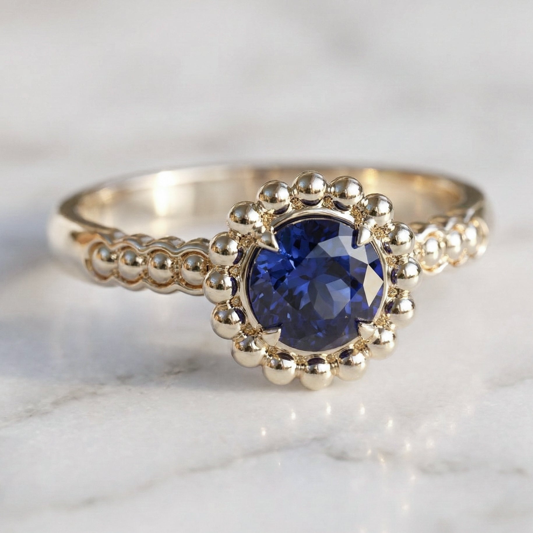 A round medium blue sapphire engagement ring with a halo of gold spheres set in a gold band.