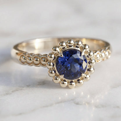 A round medium blue sapphire engagement ring with a halo of gold spheres set in a gold band.