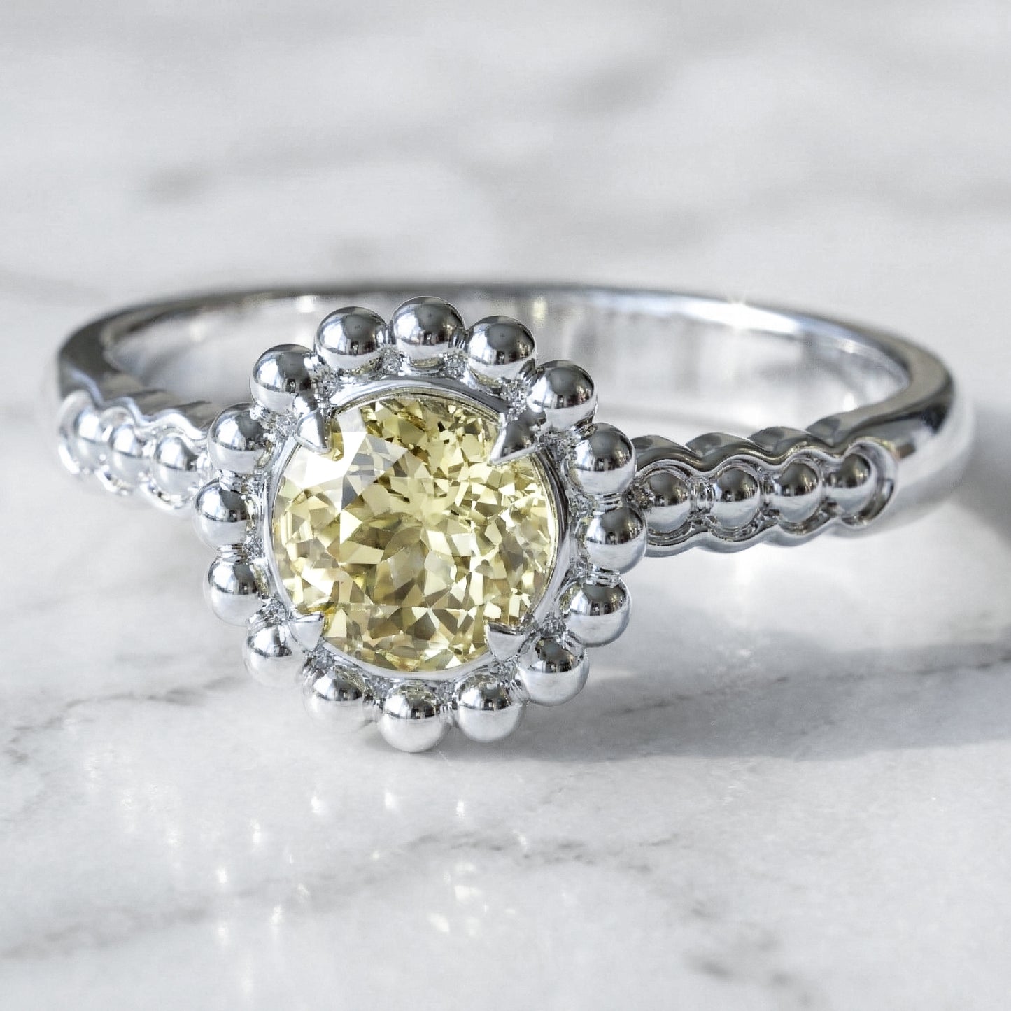 A round light yellow sapphire engagement ring with a halo of gold spheres set in a white gold band.