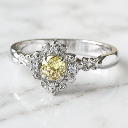 A round engagement ring with a central light yellow sapphire, flanked by four small lab-grown diamonds, set in a gold filigree setting.
