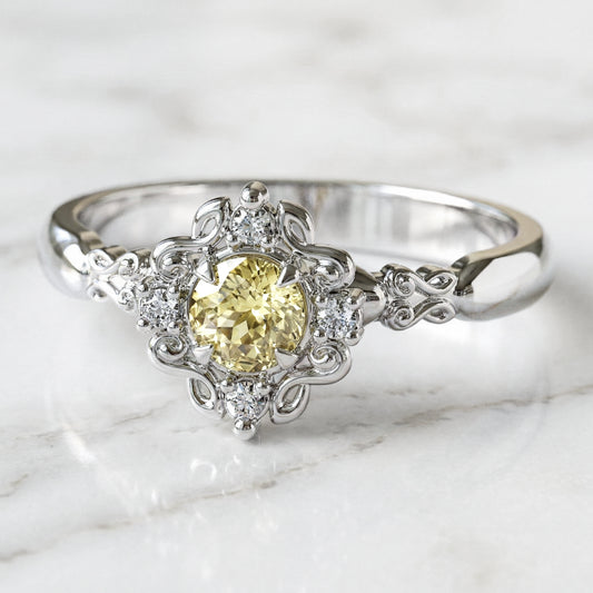 A round engagement ring with a central light yellow sapphire, flanked by four small lab-grown diamonds, set in a gold filigree setting.